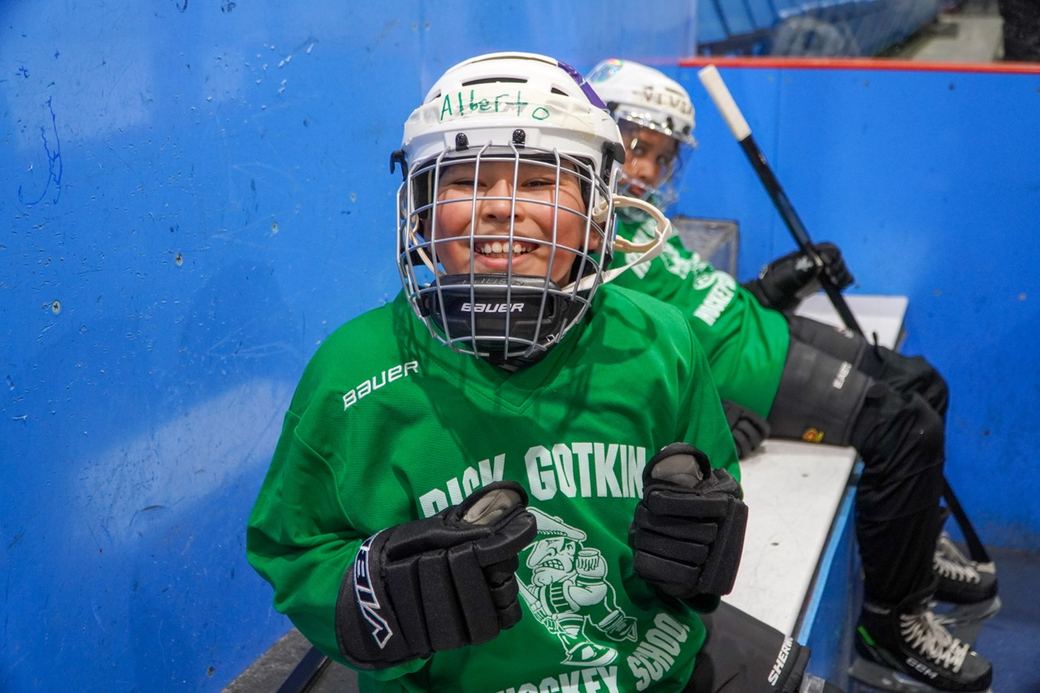 A camper in green smiling widely, stick resting on the ice