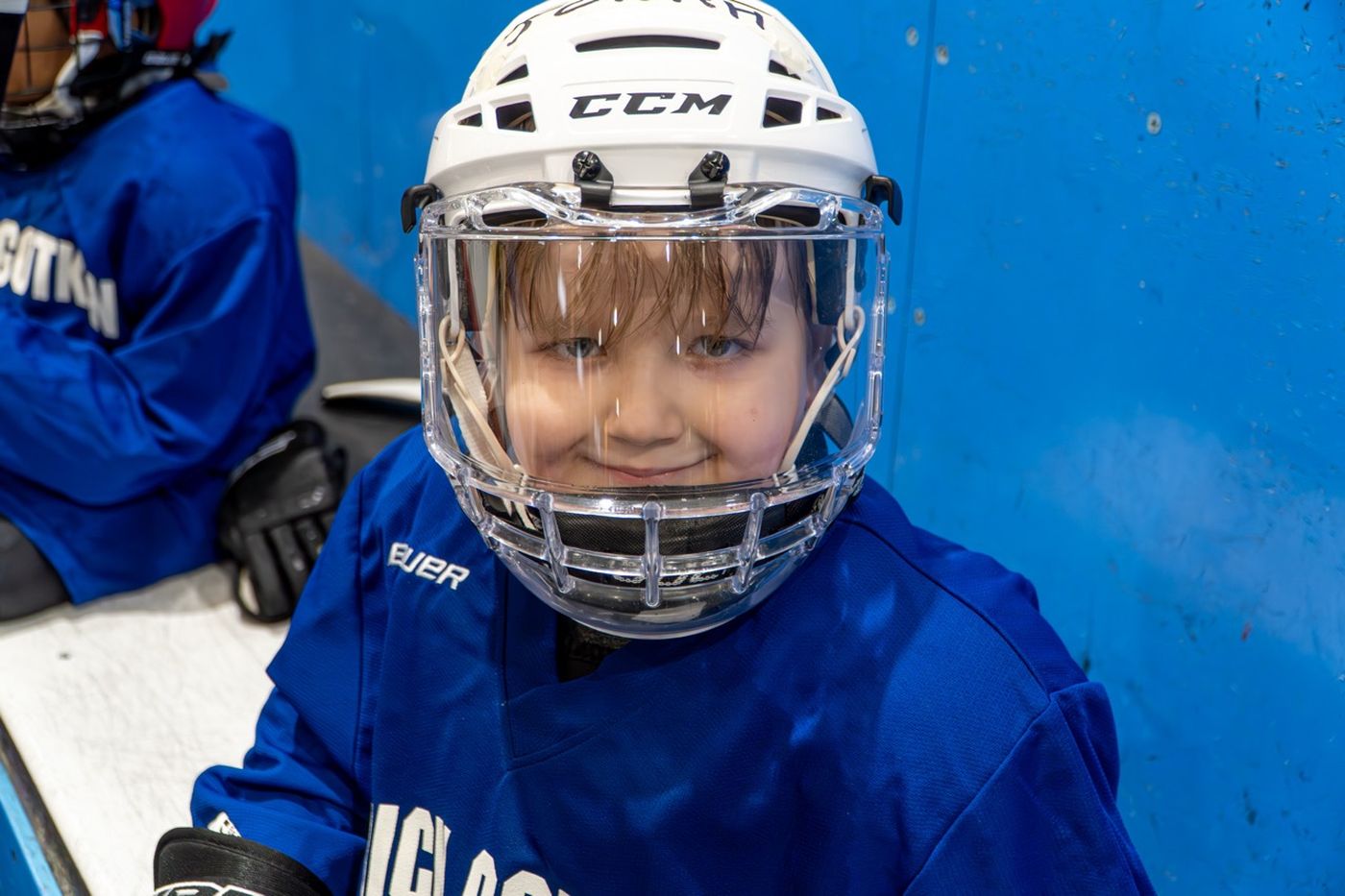 A young camper in a blue jersey and cage helmet grinning widely on the ice