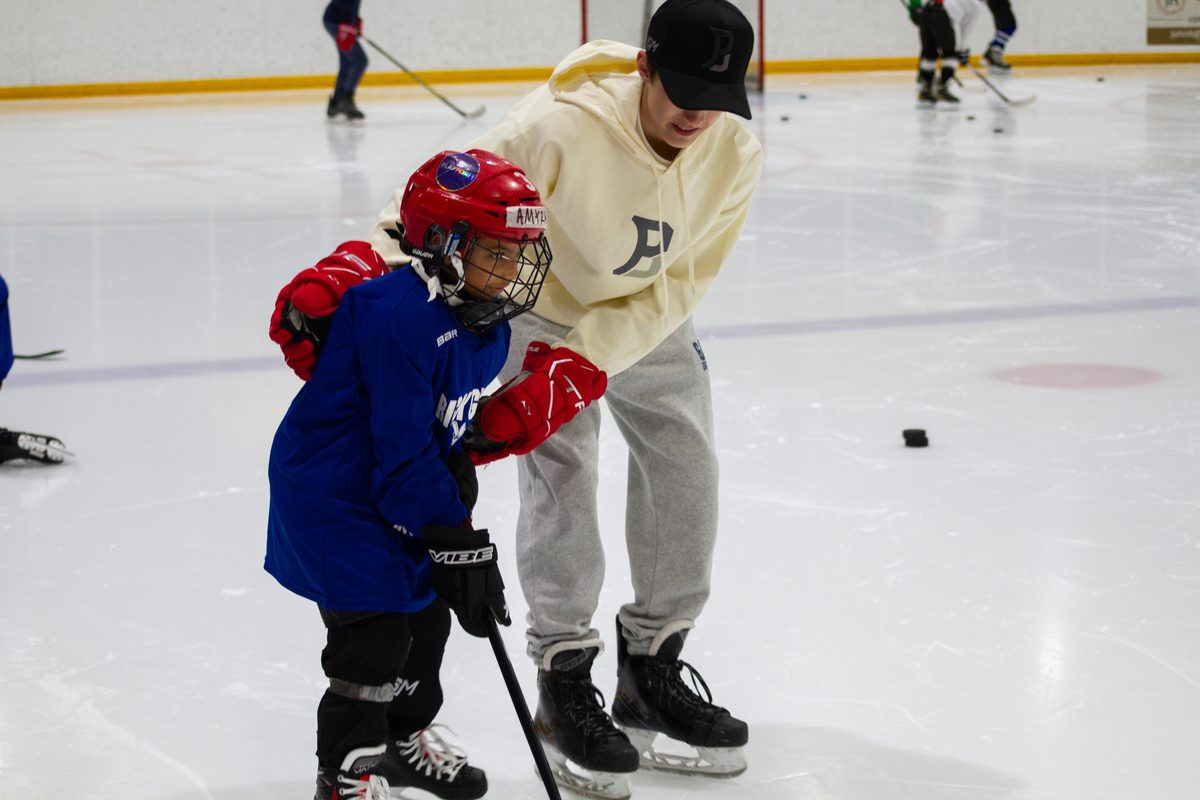 A student volunteer skating alongside a camper in a red helmet and blue jersey